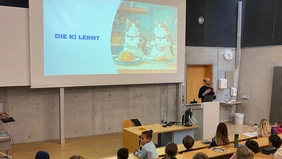 The lecturer stands at the speaker's desk in the lecture hall, behind him the presentation with two baking cats and the inscription “AI is learning”.