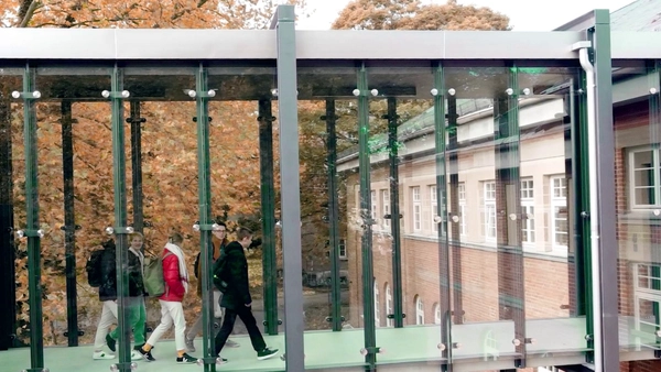 Blick von Außen in den Glasübergang des Campus Neuburg in welchem Studierende gehen.