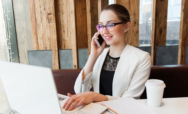 Mädchen mit Brille und Blazer sitzt am Laptop und telefoniert mit Handy.