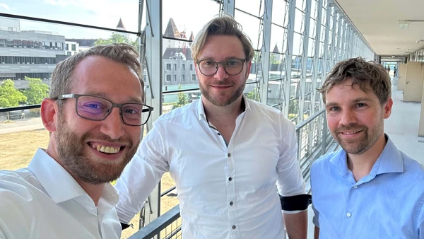 Three men standing in a classroom building of THI, with the remaining THI and the New Castle of Ingolstadt in the background. 