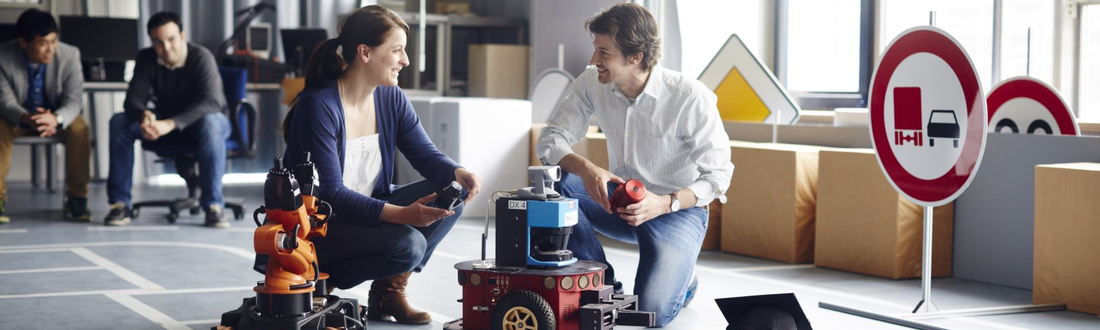 A man and a woman kneel in a test hall in front of two robots and talk