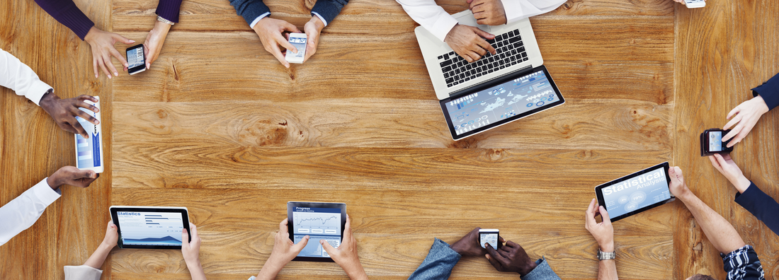 View of a wooden desk from above, showing hands, notebooks, tablets and mobile phones