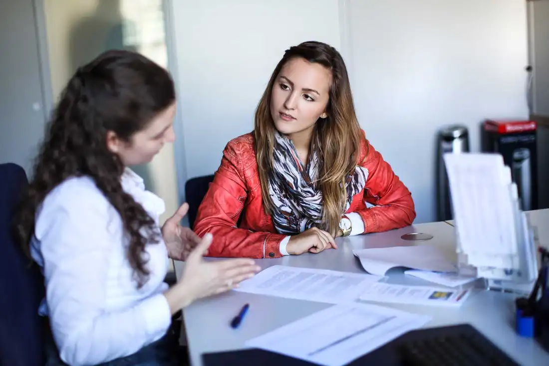 Two women sit at a table, in front of them information material which one of them explains.