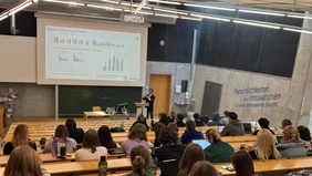 Blick von hinten in den Vorlesungssaal, vorne an der Leinwand eine Präsentation, ein Referent im Anzug am Rednerpult, die Studenten sind mit dem Rücken zu sehen