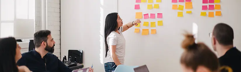 A female student stands in front of her fellow students at a whiteboard covered with Post-its 