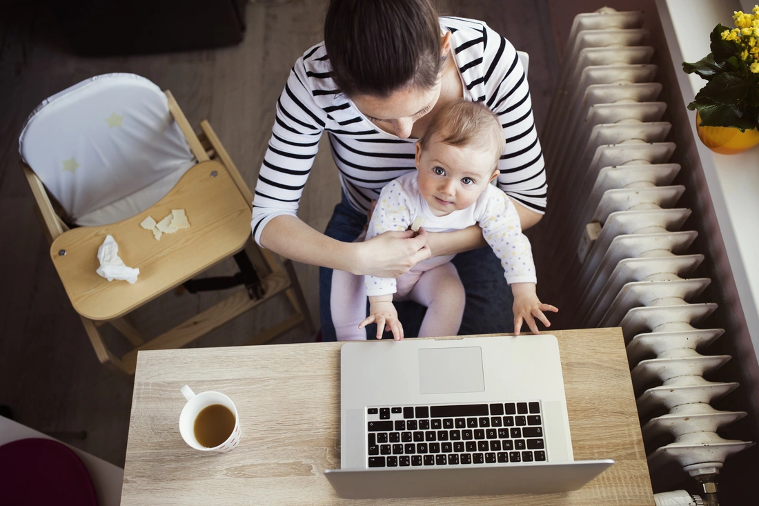 Aus der Vogelperspektive wird auf eine Frau, mit einem Baby auf dem Schoß, geschaut. Diese sitzen an einem Tisch vor einem Laptop.