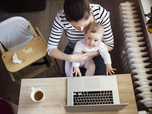 Aus der Vogelperspektive wird auf eine Frau, mit einem Baby auf dem Schoß, geschaut. Diese sitzen an einem Tisch vor einem Laptop.