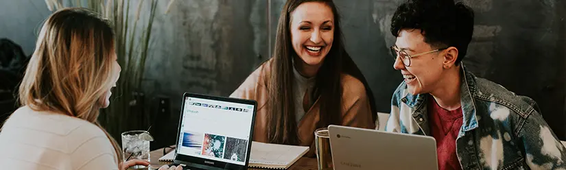 Three students sit in a café in front of their laptops and laugh