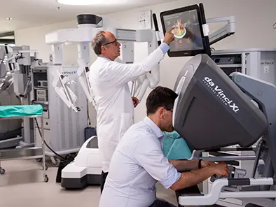 Zwei Männer in einem Raum im Klinikum. Ein Mann zeigt auf einen Bildschirm, der andere sitzt am da Vinci®-Operationssystem.