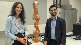 Two people look into the camera, with an orange medical robotic arm between them.