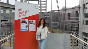 Eine Frau neben einer Stellwand, auf der zwei Plakate hängen, in der Hand ein Tablet das einen Instagram-Post zeigt, im Hintergrund die Glasfassade der THI.